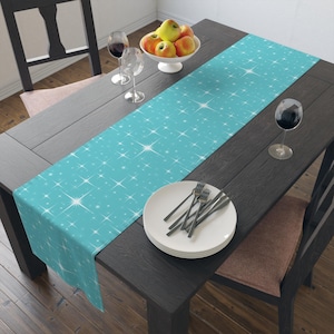 May include: A turquoise table runner with a white starburst pattern. The runner is placed on a dark wooden table, with a white bowl of fruit, wine glasses, and a plate with forks. The runner adds a decorative touch to the dining table.