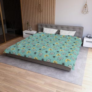 May include: A turquoise bedspread with a repeating pattern of atomic symbols, stars, and yellow half-circles. The design is in black, white, and yellow, creating a retro, mid-century modern aesthetic. The bedspread is on a gray bed frame.