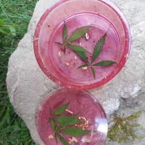 May include: Two pink resin coasters with a green marijuana leaf design and gold flakes embedded in the resin.