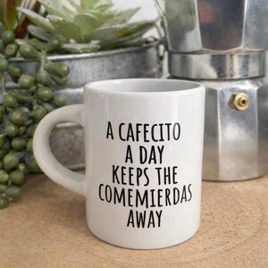 May include: A white ceramic mug with a handle and black printed text that reads A CAFECITO A DAY KEEPS THE COMEMIERDAS AWAY. The mug has a cylindrical shape with a smooth finish and is positioned in front of a metallic coffee maker and a green plant.