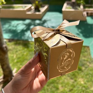 以下が含まれることがあります： 前面に花の模様と装飾的な画像が付いた、金色のギフトボックス。ボックスは、お揃いの金色のリボンで結ばれています。ボックスは人の手で持たれています。