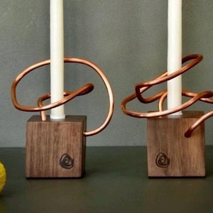 May include: Two candle holders with white candles. The holders are made of square wooden blocks with a copper wire design. The copper wire wraps around the candle. The wooden blocks have a circular design on the front.