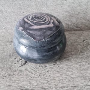 May include: A small, round, dark grey trinket box with a rose design on the lid. The rose is depicted in shades of grey and white. The box has a slightly rough texture and sits on a wooden surface.