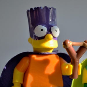 May include: A Lego figure of Bart Simpson in a superhero costume. The figure has a purple mask and cape, an orange shirt, blue shorts, and a slingshot. The figure is yellow with white eyes.
