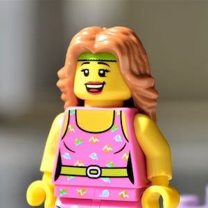 May include: A Lego minifigure of a woman with light brown hair, wearing a pink outfit with a pattern of blue and yellow shapes. The outfit includes a tank top and shorts, with a green belt. A purple boombox is in the background.