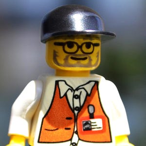 May include: A Lego minifigure of a man wearing a black cap, glasses, and an orange vest over a white shirt. The figure has a yellow face with a beard and a name tag. Black pants complete the outfit.