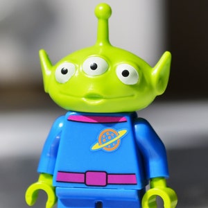 May include: A green three-eyed alien Lego minifigure wearing a blue jumpsuit with a pink belt and a planet logo on the chest.