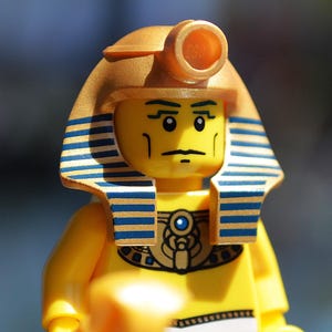 May include: A yellow Lego figurine of an Egyptian pharaoh, wearing a gold headdress and a blue and gold collar. The pharaoh holds a staff and has a stern expression. The figure's outfit includes white and blue accents.