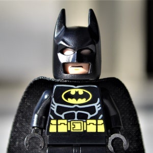 May include: A black Lego Batman figure with a black cape, a yellow utility belt, and a chest emblem featuring a yellow bat symbol. The figure has a black cowl with pointed ears and a stern expression. The figure is standing upright.