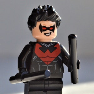 May include: A Lego figurine of a superhero character, featuring a black and red costume with a mask. The figure is holding two black batons and has a smiling expression. The character's hair is black.
