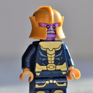 Könnte beinhalten: Eine blaue und goldene Lego-Minifigur von Thanos, einem Superschurken aus dem Marvel-Universum. Die Figur hat ein violettes Gesicht mit roten Augen und einen goldenen Helm.