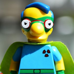 Może przedstawiać: Figurka Lego superbohatera z żółtą głową, niebieskimi włosami i okularami w zielonych oprawkach. Figurka nosi niebiesko-żółty strój z zieloną peleryną i czerwoną puszką "Buzz Cola". Figurka ma symbol promieniowania na klatce piersiowej.