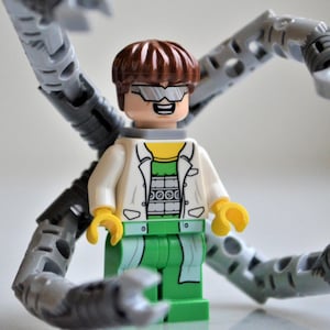 May include: A Lego figure of a character with brown hair, glasses, and a white lab coat over a yellow shirt and green pants. The figure has yellow hands and is surrounded by four gray mechanical arms.