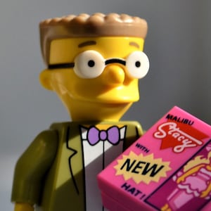 May include: A yellow Lego minifigure with brown hair wearing glasses and a green suit with a purple bow tie. The minifigure is holding a pink box with the text "Malibu Stacy with NEW HAT!"