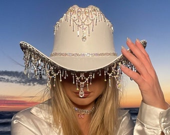 May include: A white cowboy hat with a large, ornate rhinestone design. The hat has a wide brim and a tall crown. The rhinestones are arranged in a pattern of cascading teardrops and geometric shapes.