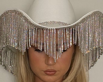 May include: A white cowboy hat with a wide brim and a band of rhinestones. The hat is decorated with long, dangling strands of rhinestones.