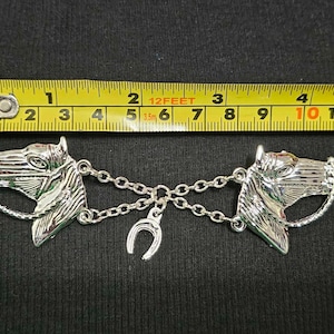 May include: Silver horse head brooch with a chain connecting two horse heads. The chain has a horseshoe charm in the center.