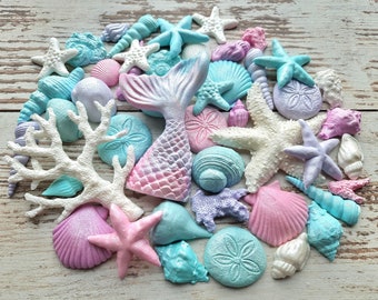 May include: A collection of pastel-colored seashells, starfish, and coral. The shells are in shades of pink, blue, and purple. There is a mermaid tail made of fondant in the center of the image.