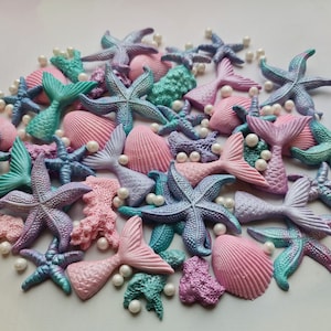 Puede incluir: Una colección de decoraciones para pasteles de temática de sirena en colores pastel, que incluyen conchas marinas, estrellas de mar y colas de sirena. Las decoraciones están hechas de fondant y son perfectas para agregar un toque caprichoso a cualquier pastel.