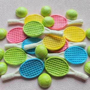 K&ouml;nnte beinhalten: Eine Sammlung bunter Tennisschl&auml;ger und Tennisb&auml;lle. Die Schl&auml;ger sind in den Farbt&ouml;nen Rosa, Gelb, Gr&uuml;n und Blau gehalten und haben wei&szlig;e Griffe. Die Tennisb&auml;lle sind hellgr&uuml;n. Die Artikel sind auf einer rosa strukturierten Oberfl&auml;che angeordnet.