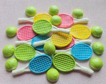 May include: A collection of colorful tennis rackets and tennis balls. The rackets are in shades of pink, yellow, green, and blue, with white handles. The tennis balls are a bright green color. The items are arranged on a pink textured surface.