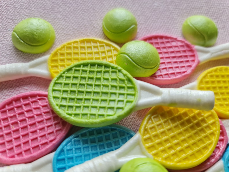 K&ouml;nnte beinhalten: Eine Sammlung bunter Tennisschl&auml;ger und Tennisb&auml;lle. Die Schl&auml;ger sind in Gelb-, Rosa-, Gr&uuml;n- und Blaut&ouml;nen gehalten und haben wei&szlig;e Griffe. Die Tennisb&auml;lle sind hellgr&uuml;n. Die Artikel sind auf einer hellrosa Oberfl&auml;che angeordnet.