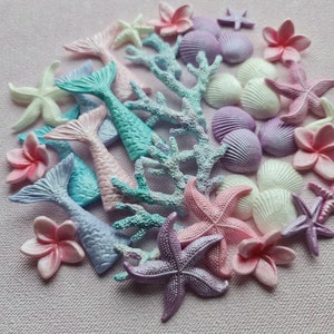 Op de afbeelding: Een verzameling pastelkleurige zeemeerminstaart- en zeester-taarttoppers, samen met schelpen en koraal. De decoraties zijn perfect voor een zeemeermin-themafeest of taart.