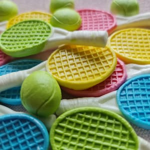 K&ouml;nnte beinhalten: Eine Sammlung bunter Tennisracket- und Ball-f&ouml;rmiger Dekorationen. Die Schl&auml;ger sind in Gr&uuml;n-, Gelb-, Rosa- und Blaut&ouml;nen gehalten, mit wei&szlig;en Griffen. Die Tennisb&auml;lle sind gr&uuml;n. Die Artikel sind auf einer hellrosa Oberfl&auml;che angeordnet.