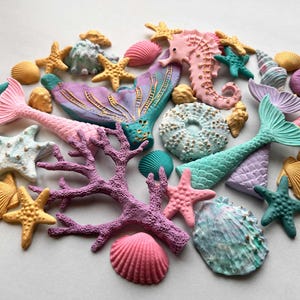 Puede incluir: Una colección de elementos decorativos con temática marina. Incluye estrellas de mar, conchas, colas de sirena, un caballito de mar y coral en tonos rosa, turquesa, amarillo y morado. Ideal para manualidades o decoración temática.
