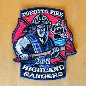 May include: An embroidered patch featuring a firefighter holding an axe. The patch is red, white and blue and reads "Toronto Fire" at the top, "215 Highland Rangers" at the bottom, and "11" on the firefighter's helmet.