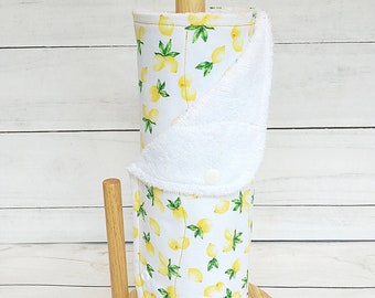 May include: A white paper towel holder with a lemon print design. The holder is made of fabric and has a white terry cloth lining. The holder is on a wooden stand.