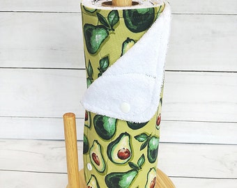 May include: A green and white reusable paper towel roll with an avocado print. The roll is on a wooden stand and has a white terry cloth cover.