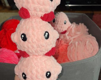 May include: A stack of three handmade pink crochet octopus toys with red accents and black eyes. The toys are stacked on top of each other, with a blue charm attached to the bottom one. Other pink yarn and octopus toys are in a gray bin.