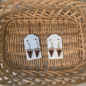 May include: Two pairs of dangle earrings featuring a brown and tan pie slice design. The earrings are displayed on white card stock.