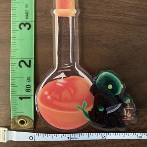 May include: A clear glass bottle keychain with a gold star charm. The bottle is filled with a glittery orange liquid and has a green plant growing out of it. A small black and green creature with a lantern is attached to the bottle.
