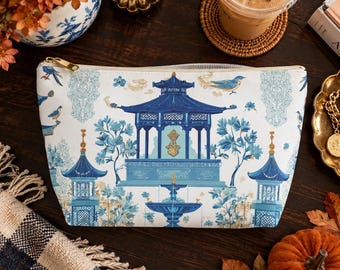 Puede incluir: Un estuche blanco con cremallera que presenta un estampado de estilo chinoiserie en azul y dorado con una pagoda central, pabellones laterales, una fuente y pájaros. El estuche tiene un cierre de cremallera de metal dorado.