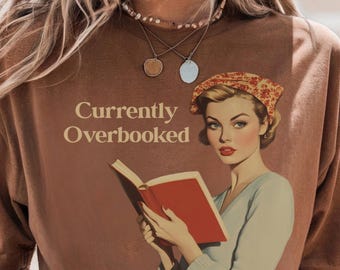 Op de afbeelding: Bruin T-shirt met de tekst "Currently Overbooked" en een vintage illustratie van een vrouw die een boek leest. De vrouw draagt een hoofddoek en een lichtblauwe top. Het boek heeft een rode kaft.