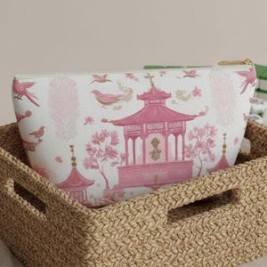 May include: A white cosmetic bag with a pink pagoda and bird design, set inside a woven basket. The bag features a gold zipper and a repeating pattern of pink birds and floral elements. The basket is made of a light brown woven material.
