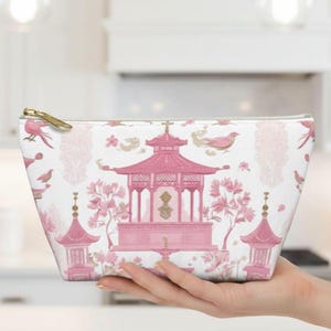May include: A white cosmetic bag with a pink Asian-inspired design. The pattern includes pagodas, birds, and floral elements. The bag has a gold zipper and is held in a hand.
