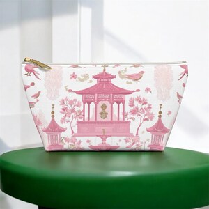 May include: A white cosmetic bag with a pink Asian-inspired design featuring pagodas, birds, and floral elements. The bag has a gold zipper and is sitting on a green surface. The bag is likely used for storing makeup or toiletries.