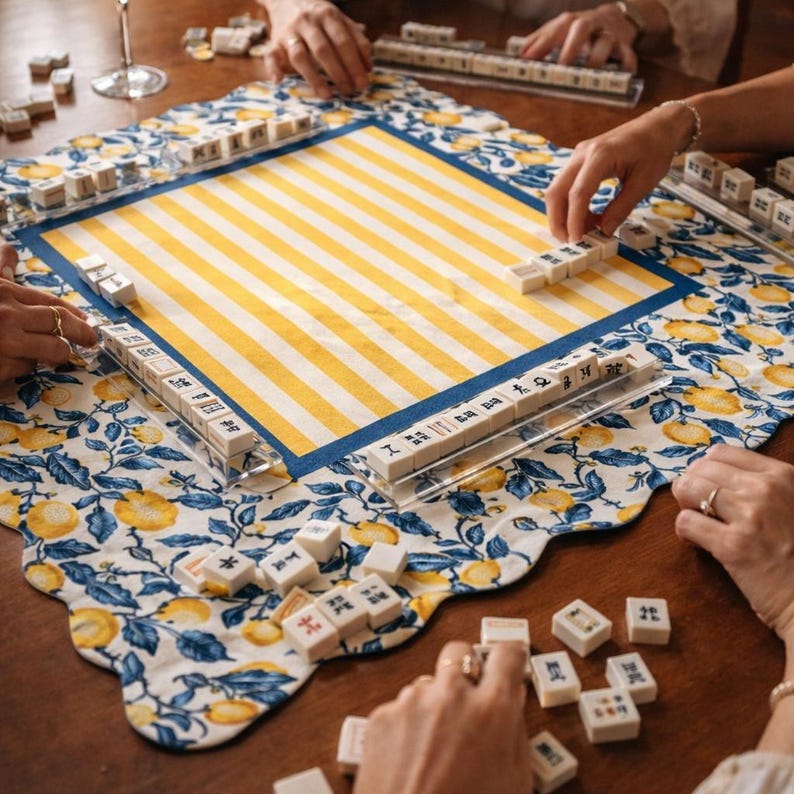 May include: A Mahjong game in progress, featuring a yellow and white striped playing surface with a blue border. The game is set on a floral patterned cloth with lemons and blue leaves. Players are arranging tiles.