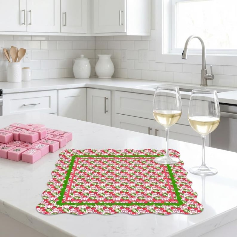 May include: A patterned placemat with a scalloped edge, featuring red strawberries on a white background, framed by a green border and a pink inner line. Two wine glasses filled with white wine are on the counter.