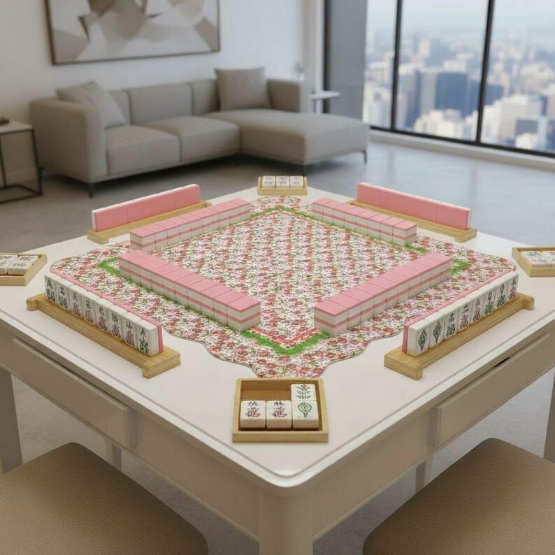 May include: A white square Mahjong table with a floral patterned playing surface. Pink and white tiles are arranged on wooden racks around the table. The table is set in a modern living room with a large window.