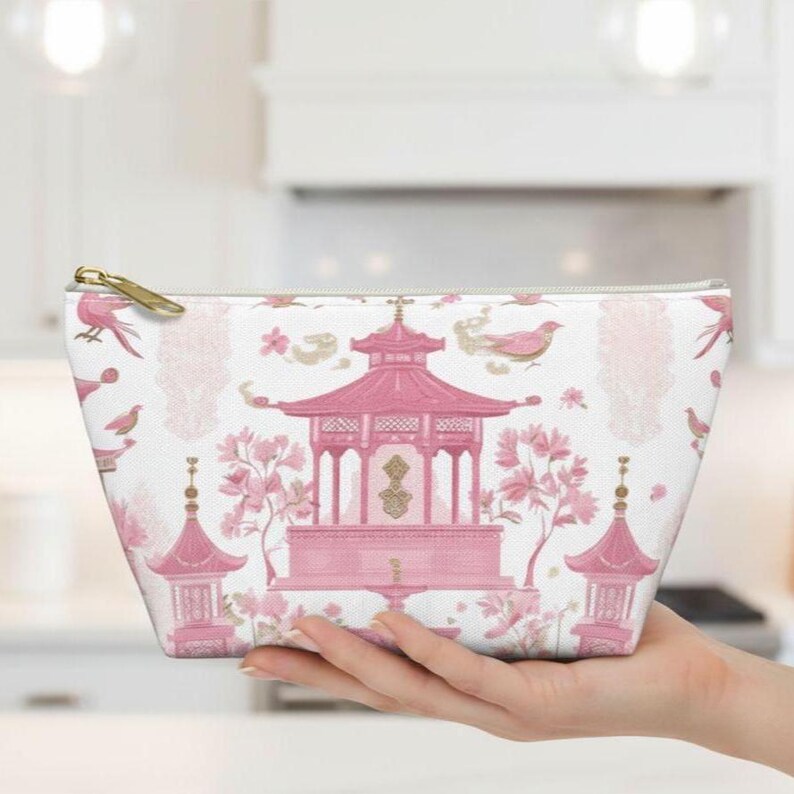 May include: A white cosmetic bag with a pink Asian-inspired design. The pattern includes pagodas, birds, and floral elements. The bag has a gold zipper and is held in a hand.