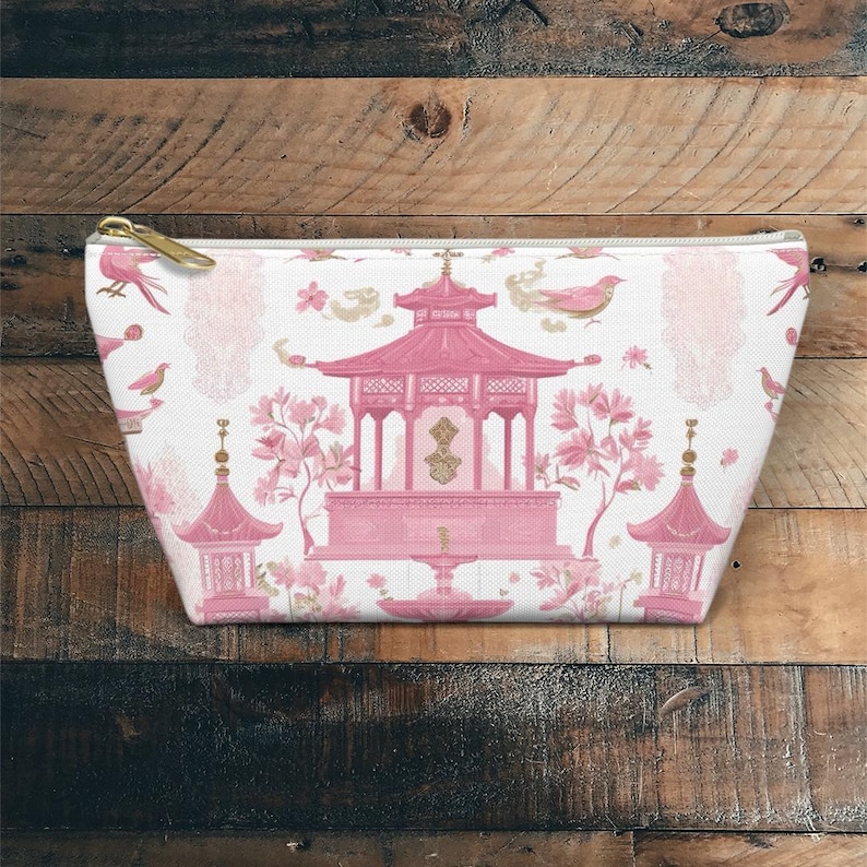 May include: A white cosmetic bag with a pink Asian-inspired design. The design features a pagoda, birds, and floral elements. The bag has a gold zipper and is made of a soft, textured fabric.