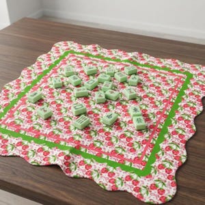 May include: A Mahjong set on a floral mat with a strawberry pattern. The mat has green and pink borders. The Mahjong tiles are light green with black symbols. The mat is on a wooden table.