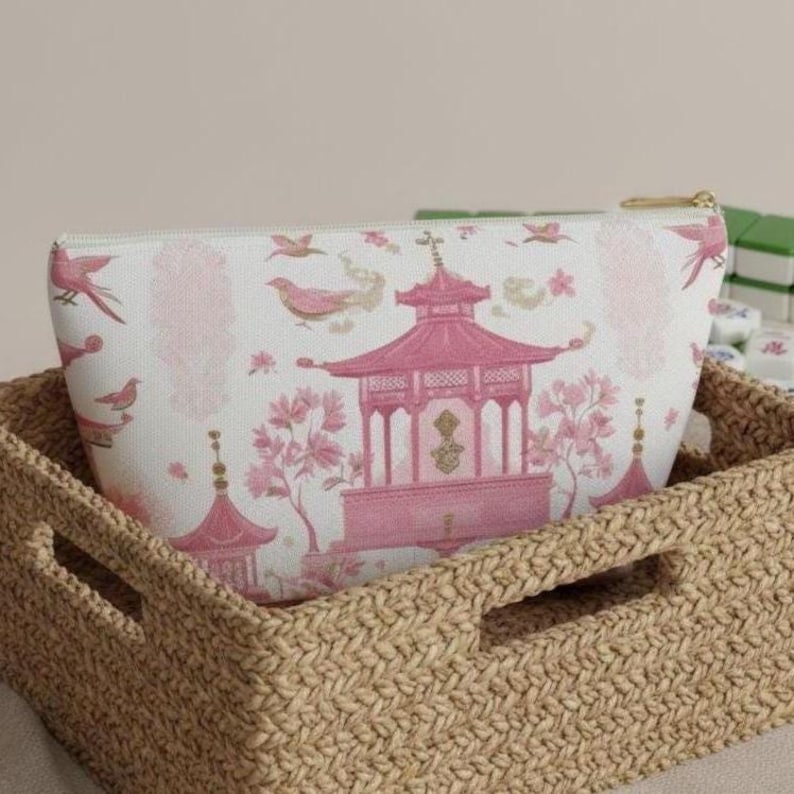 May include: A white cosmetic bag with a pink pagoda and bird design, set inside a woven basket. The bag features a gold zipper and a repeating pattern of pink birds and floral elements. The basket is made of a light brown woven material.