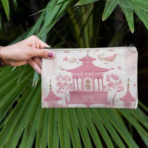 May include: A rectangular cosmetic bag with a pink pagoda design. The bag features a light pink and white background with a repeating pattern. The pagoda is a darker shade of pink, with floral and bird accents.