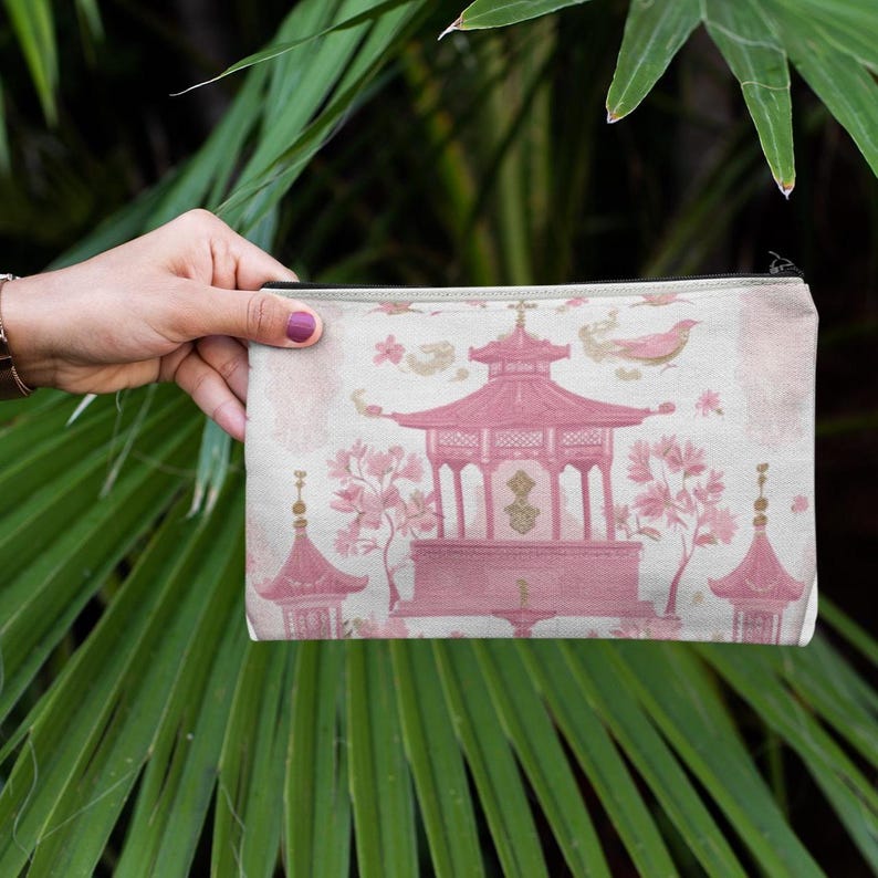 May include: A rectangular cosmetic bag with a pink pagoda design. The bag features a light pink and white background with a repeating pattern. The pagoda is a darker shade of pink, with floral and bird accents.