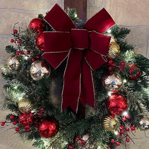 May include: A Christmas wreath with a large burgundy velvet bow, gold and red ornaments, and sprigs of red berries.